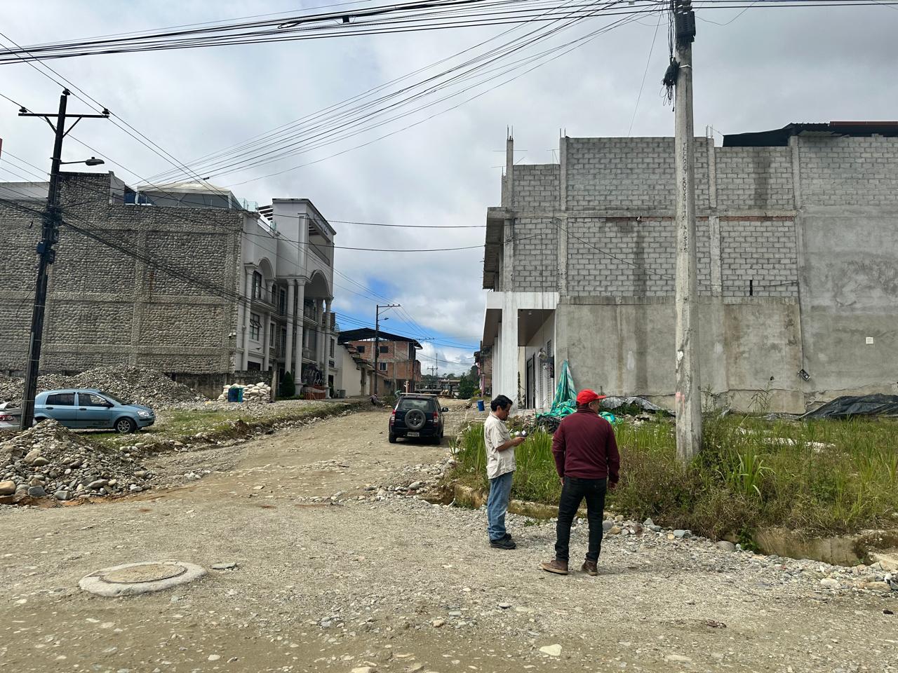 Dos personas en una calle de tierra con maleza y viviendas en Yantzaza, evaluando el entorno urbano por riesgo de dengue