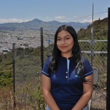Retrato de Hilary Madeley Calva Camacho vistiendo el uniforme del Grupo de Investigación en Hidroinformática, con equipos de monitoreo ambiental y una vista panorámica de Loja al fondo.