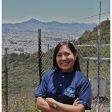 Retrato de Marjorie Cristina Díaz López sonriendo con los brazos cruzados, vistiendo el uniforme del Grupo de Investigación en Hidroinformática, con una vista panorámica de Loja y una estación meteorológica al fondo.