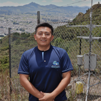 Retrato de Edison Stalin Salinas Nalvay usando el uniforme del Grupo de Investigación en Hidroinformática, junto a una estación de monitoreo ambiental con una vista panorámica de la ciudad de Loja al fondo.