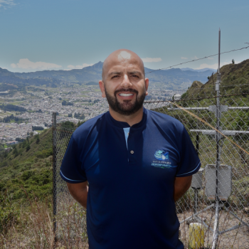 Retrato de Luis Felipe Duque sonriendo, vistiendo una camiseta del Grupo de Investigación en Hidroinformática, con una vista panorámica de la ciudad de Loja y equipos de monitoreo ambiental al fondo.