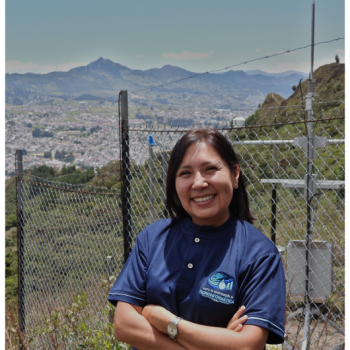 Retrato de Marjorie Cristina Díaz López sonriendo con los brazos cruzados, vistiendo el uniforme del Grupo de Investigación en Hidroinformática, con una vista panorámica de Loja y una estación meteorológica al fondo.